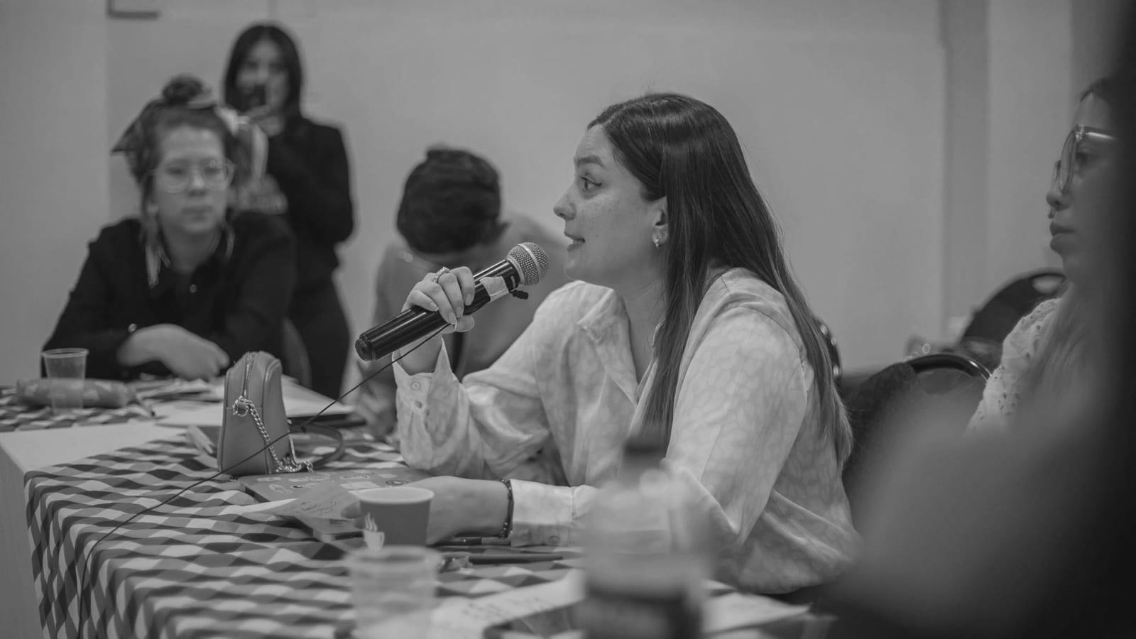 A practitioner speaking into a microphone at a panel table while colleagues listen.